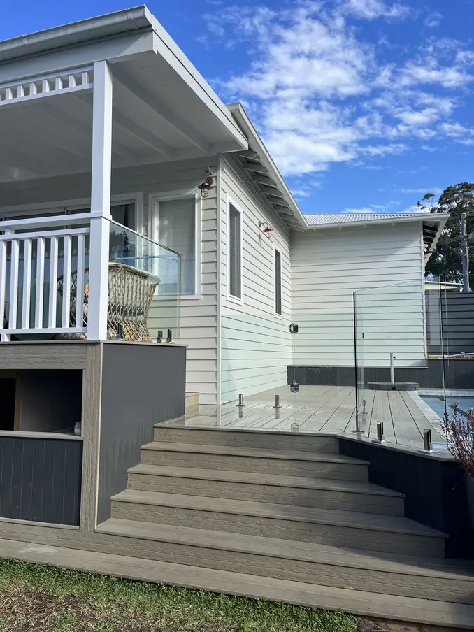 Wide deck steps leading up to a covered verandah with white timber balustrade, beside a pool with frameless glass fencing