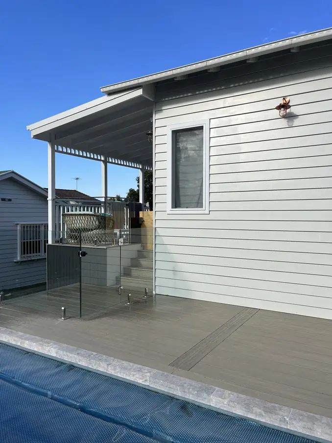 Grey deck running to the edge of a pool, with frameless glass fencing and a weatherboard house behind